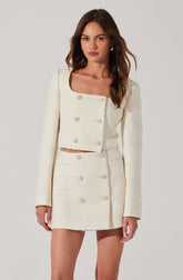 A woman is wearing a cream-colored, textured two-piece outfit consisting of a fitted, long-sleeved crop top with gold buttons and a matching high-waisted mini skirt also with gold buttons. The crop top has a square neckline and a boxy silhouette.